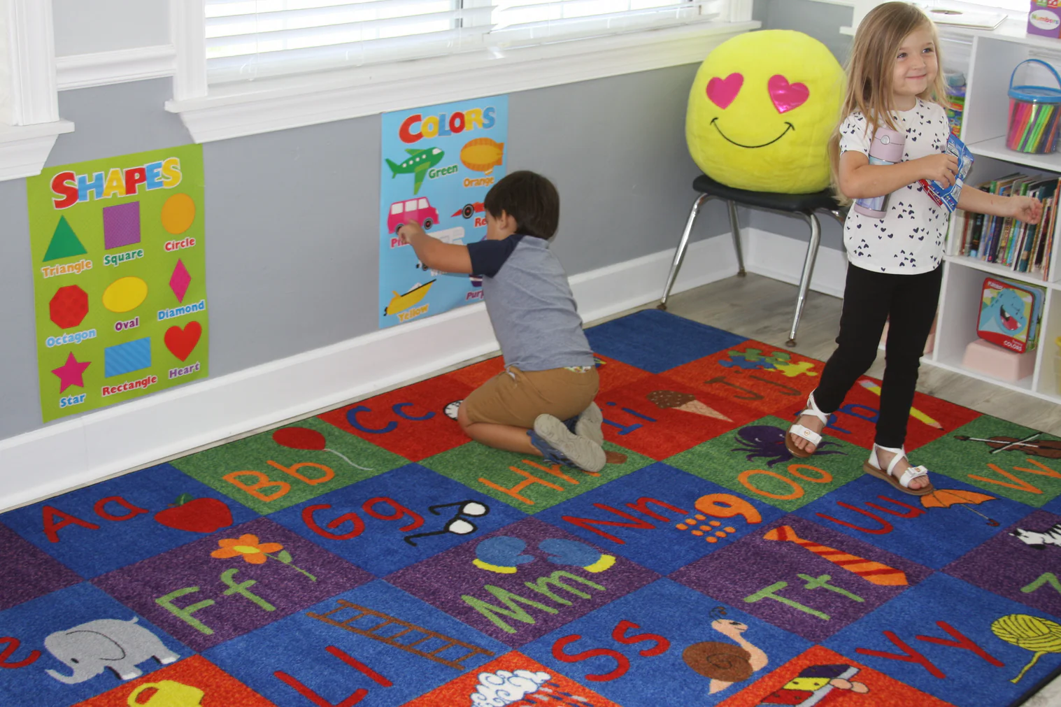 Children interacting with a colorful educational rug featuring letters, numbers, and phonics visuals in a bright preschool classroom, promoting hands-on literacy and numeracy learning.